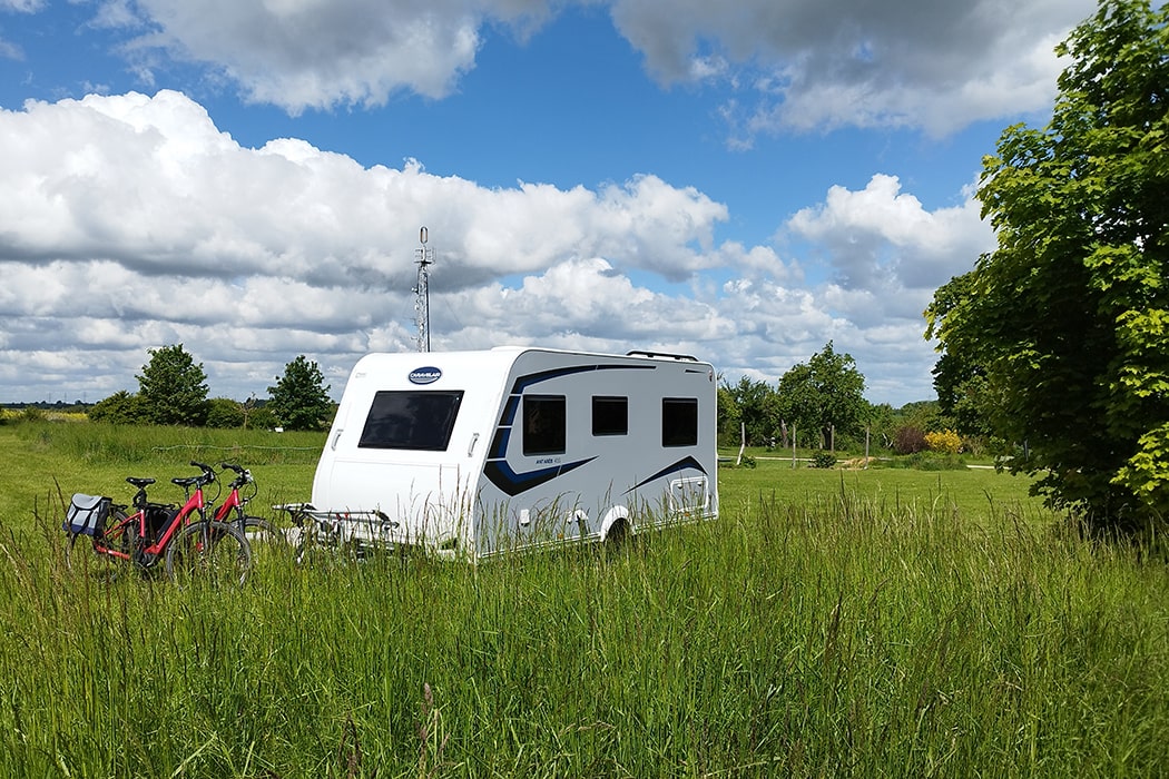 Weißer Wohnwagen mit zwei Fahrrädern davor auf einer Wiese mit hohen Gräsern auf dem Campingplatz MiO in Ottendorf bei Chemnitz. Im Hintergrund Bäume und ein Mobilfunkmast.