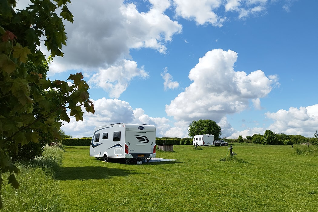 Zwei Wohnwagen auf einer weitläufigen Wiese auf dem Campingplatz MiO in Ottendorf bei Chemnitz. Im Vordergrund ein Baum mit grünem Laub, darüber ein Himmel mit Wolken.