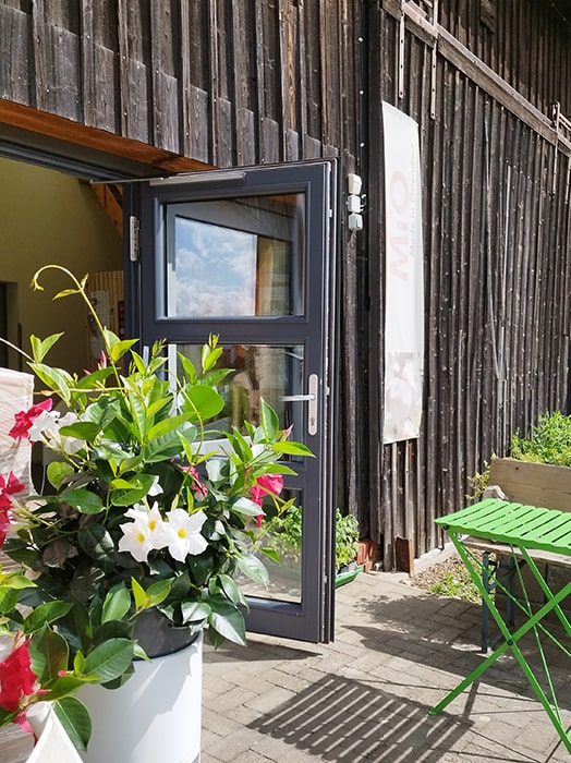 Offene Glastür an einem Holzgebäude auf dem Campingplatz MiO in Ottendorf bei Chemnitz. Daneben ein Blumenkübel mit roten und weißen Blüten sowie ein grüner Klapptisch mit Bank.
