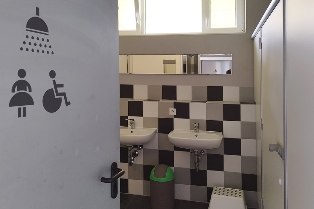 Eingang zu einem Sanitärraum mit zwei Waschbecken, Spiegel, Mülleimer und Tritthocker auf dem Campingplatz MiO in Ottendorf bei Chemnitz. An der geöffneten Tür sind Piktogramme für Dusche, Frauen und Rollstuhlnutzer zu sehen.