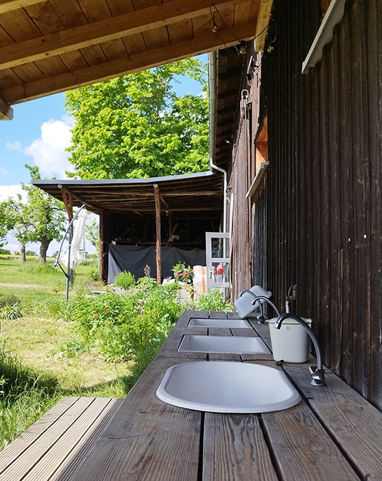 Mehrere Spülbecken mit Wasserhähnen an einer Holztheke im Außenbereich eines Gebäudes auf dem Campingplatz MiO in Ottendorf bei Chemnitz. Im Hintergrund eine überdachte Sitzfläche und Bäume.
