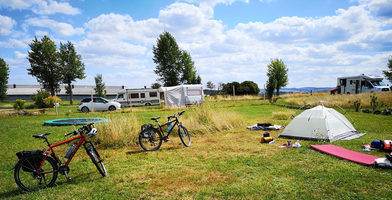 Zelt, Fahrräder, Campingausrüstung und Fahrzeuge auf einer offenen Wiese auf dem Campingplatz MiO in Ottendorf bei Chemnitz. Im Hintergrund Bäume, Wohnmobile und ein weiter Blick über die Landschaft.