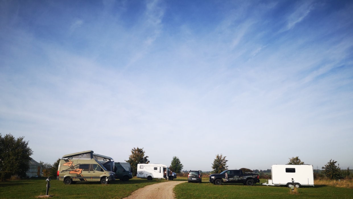 Mehrere Camper und Wohnmobile stehen auf einer Wiese unter blauem Himmel mit leichten Wolken